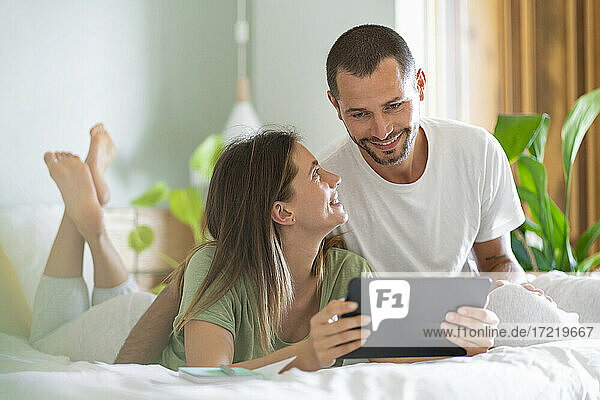 Young woman with digital tablet looking at husband sitting on bed at home