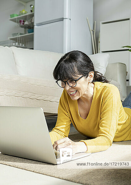 Lächelnde Frau  die einen Laptop benutzt  während sie zu Hause auf dem Boden liegt