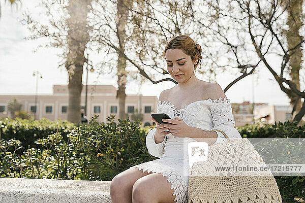 Junge Frau im weißen Kleid  die im Park sitzend ein Mobiltelefon benutzt