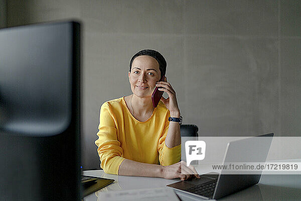 Zufriedene Unternehmerin mit Laptop  die am Schreibtisch in einem modernen Büro mit ihrem Mobiltelefon spricht