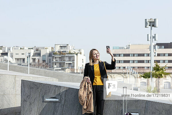 Lächelnde Geschäftsfrau mit Jacke und Koffer  die ein Selfie mit ihrem Mobiltelefon in der Stadt macht