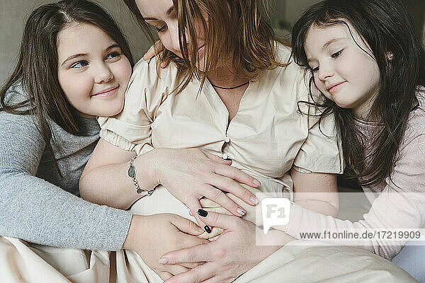 Aufgeregte schwangere Mutter und Töchter mit Händen auf dem Bauch sitzen zu Hause