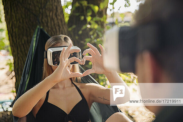 Paar entspannt sich in Hängematte am See  mit VR-Brille  Frau zeigt herzförmigen Fingerrahmen