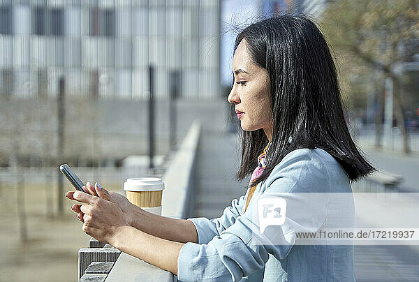 Geschäftsfrau  die eine Textnachricht über ihr Smartphone sendet  während sie sich auf ein Geländer stützt