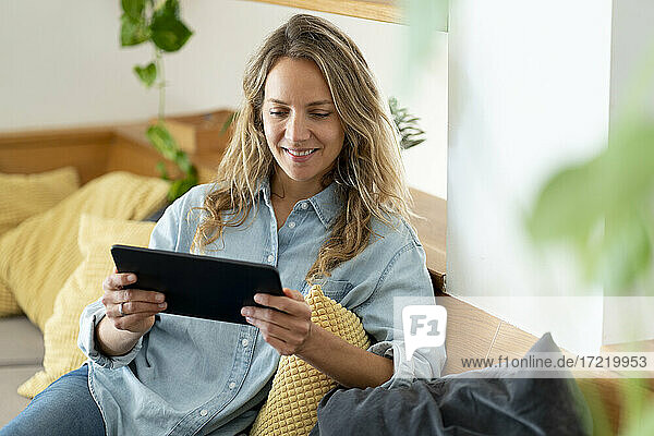 Lächelnde Frau  die ein digitales Tablet benutzt  während sie zu Hause auf der Couch im Wohnzimmer sitzt