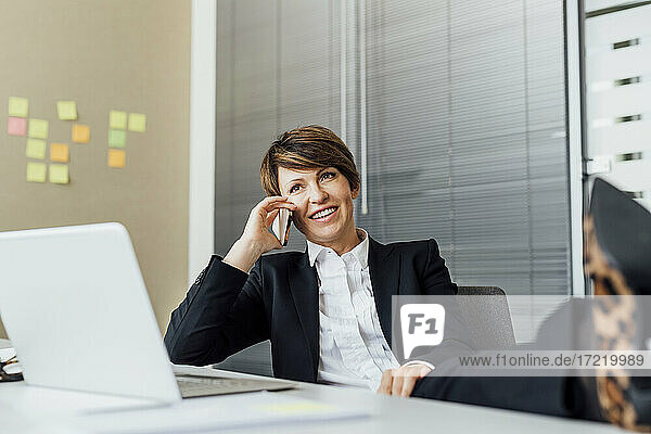 Glückliche Geschäftsfrau  die wegschaut  während sie am Schreibtisch mit ihrem Smartphone telefoniert