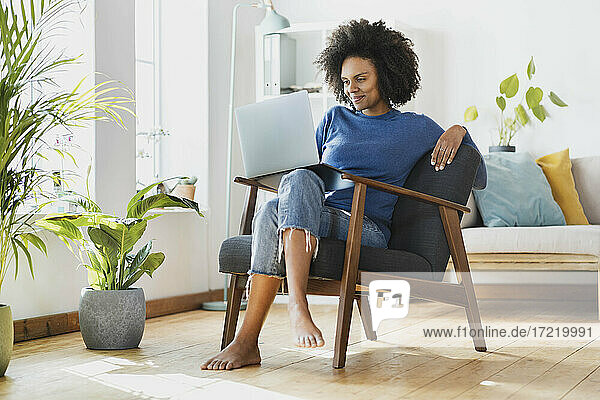 Lächelnde Frau  die einen Laptop benutzt  während sie zu Hause auf einem Sessel sitzt