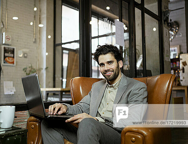 Lächelnde männliche Fachkraft auf einem Sessel sitzend mit Laptop in einem beleuchteten Café