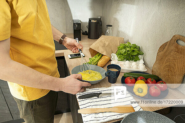 Mann rührt Eier in Schüssel mit Schneebesen auf Küchentisch zu Hause