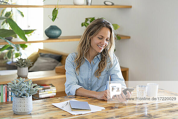 Smiling blond female entrepreneur surfing net through smart phone while sitting at table