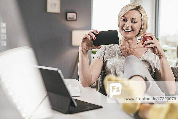 Lächelnde Frau mit Apfel  die zu Hause einen Film auf dem Mobiltelefon ansieht