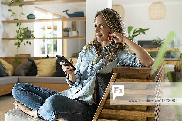 Smiling woman sitting with mobile phone on couch while looking away