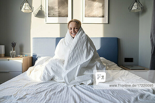 Lächelnde Frau  eingewickelt in eine Decke  sitzt zu Hause auf dem Bett