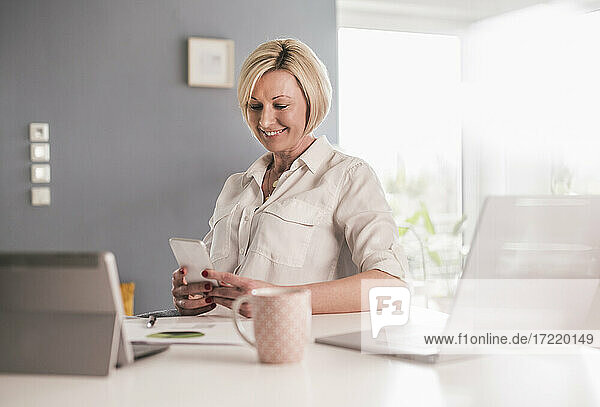 Lächelnde weibliche Fachkraft  die im Büro zu Hause an einem Smartphone arbeitet