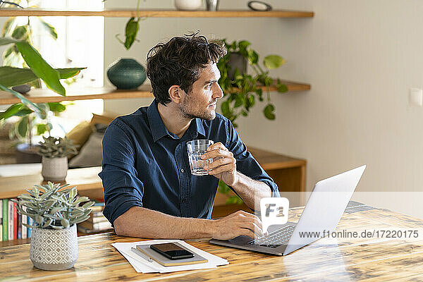 Nachdenklicher männlicher Freiberufler trinkt Wasser  während er mit seinem Laptop am Tisch sitzt
