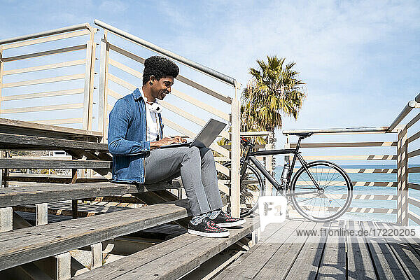Smiling man using laptop while sitting on steps at promenade by bicycle during sunny day
