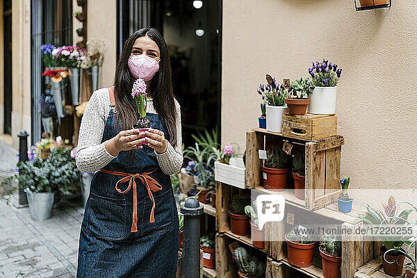 Young female florist with protective face mask holding potted plant while working at flower shop