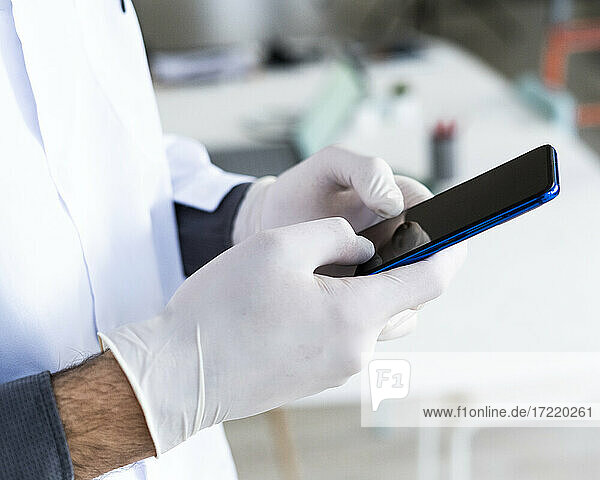 Männlicher medizinischer Angestellter mit chirurgischen Handschuhen  der im Krankenhaus eine Textnachricht über sein Mobiltelefon sendet