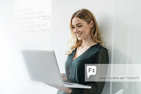Lächelnde Geschäftsfrau  die einen Laptop benutzt und sich im Büro an die Wand lehnt
