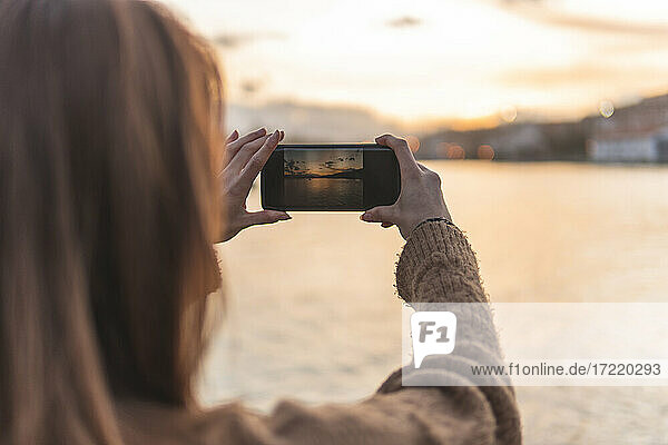 Spanien  Kantabrien  San Vicente de la Barquera  Junge Frau fotografiert Meeresküste bei Sonnenuntergang