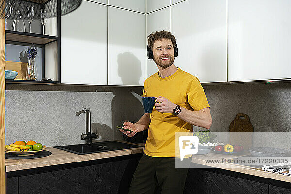 Mann hört Musik und hält Handy und Kaffeetasse in der Küche zu Hause