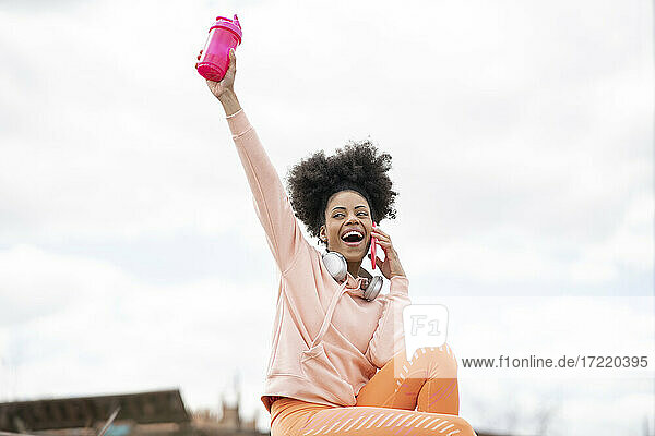 Fröhliche Frau  die einen Shaker hält  während sie mit einem Smartphone telefoniert