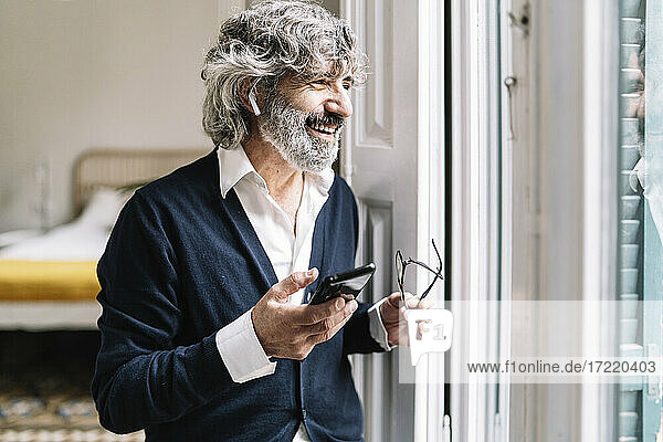Lächelnder älterer Mann mit In-Ear-Kopfhörern  der in der Nähe des Fensters zu Hause mit seinem Smartphone telefoniert