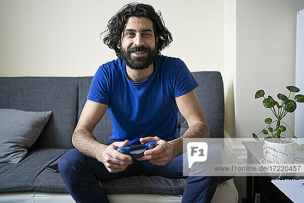 Lächelnder Mann mit Gamecontroller  der zu Hause auf dem Sofa sitzt