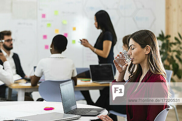 Junge Geschäftsfrau trinkt Wasser  während sie im Büro am Laptop sitzt