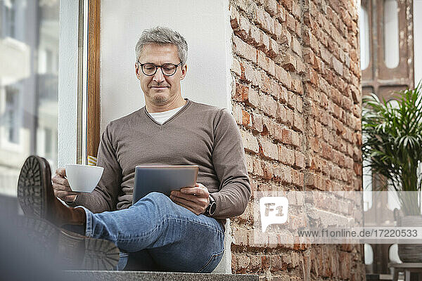 Mann hält Kaffeetasse  während er ein digitales Tablet zu Hause benutzt