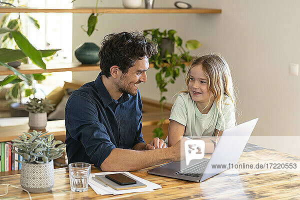 Smiling father and daughter looking at each other in front of laptop at home