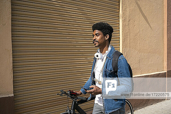 Afro-Mann mit Fahrrad hält Smartphone an der Wand während eines sonnigen Tages