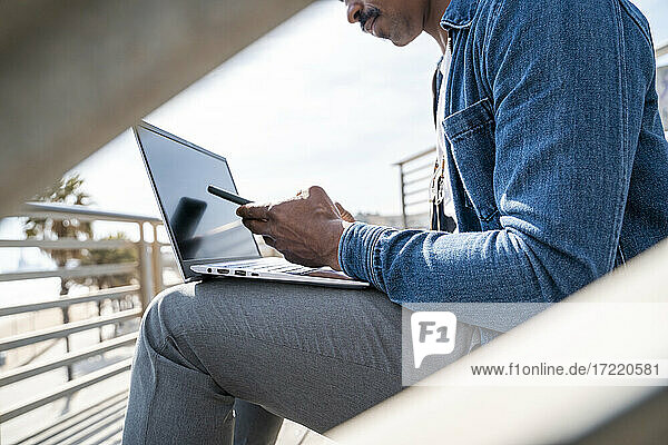 Mann benutzt sein Smartphone  während er mit seinem Laptop auf der Promenade sitzt