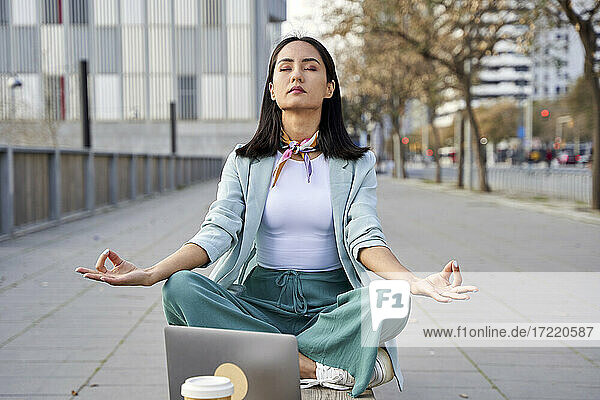 Junge Geschäftsfrau mit Laptop meditiert auf einer Bank am Fußweg