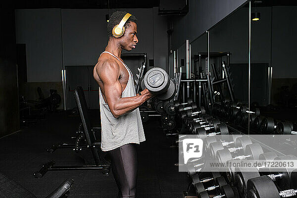 Male athlete listening music through headphones while exercising with dumbbell at gym