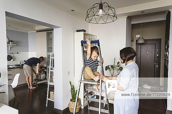 Mutter spielt mit ihrem Sohn auf der Leiter sitzend  während der Vater zu Hause in der Küche arbeitet