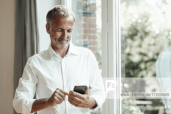 Älterer Mann mit Smartphone am Fenster stehend zu Hause