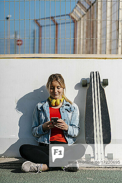 Lächelnde junge Frau mit gekreuzten Beinen  die am Longboard ein Mobiltelefon benutzt