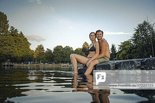 Junges Paar sitzt auf einer Badeplattform an einem See und schaut in die Ferne