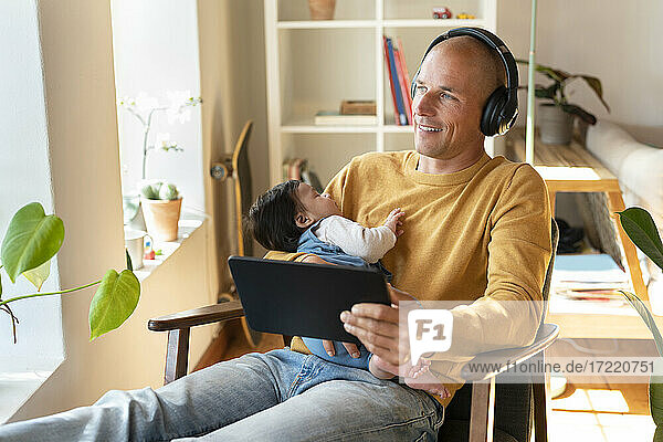 Lächelnder Vater hört Musik  während er ein schlafendes Baby und ein digitales Tablet im Wohnzimmer hält