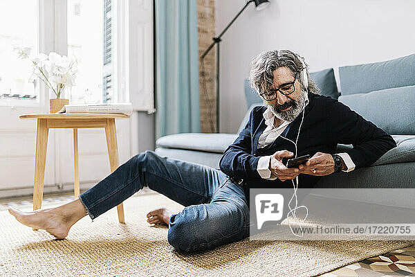 Mann benutzt Smartphone  während er zu Hause auf dem Teppich sitzt