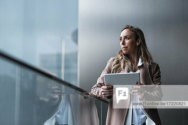 Unternehmerin träumt  während sie ein digitales Tablet vor einer grauen Wand hält