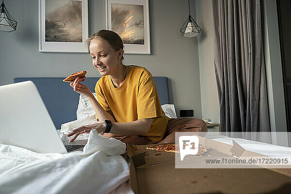 Lächelnde Frau  die ein Pizzastück hält  während sie zu Hause einen Laptop benutzt