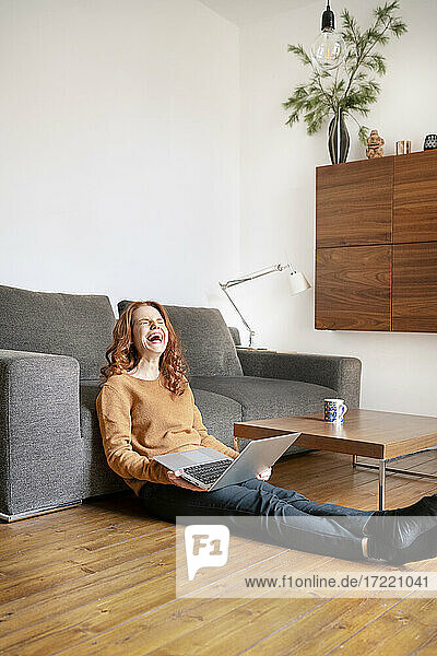 Fröhliche Frau lacht in voller Länge  während sie mit einem Laptop im Wohnzimmer zu Hause sitzt