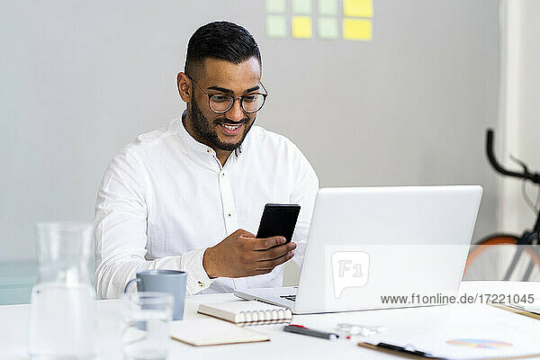 Junger Geschäftsmann  der ein Smartphone neben einem Laptop auf dem Schreibtisch im Büro benutzt