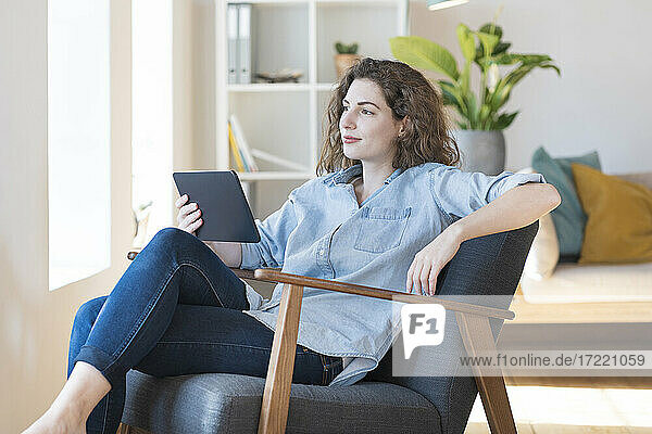 Junge Frau  die auf einem Sessel zu Hause ein digitales Tablet hält und träumt