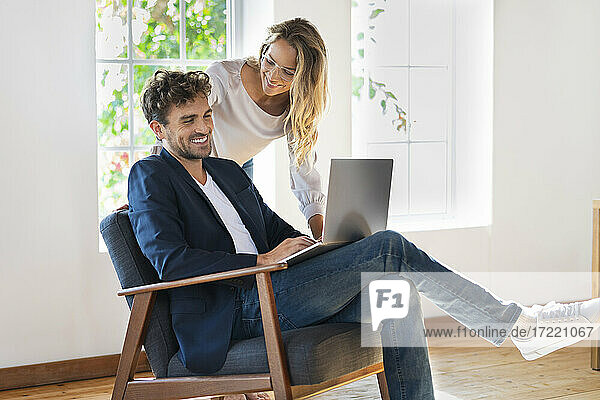 Lächelnde Freundin sieht ihren Freund an  der einen Laptop benutzt  während er zu Hause auf einem Stuhl sitzt
