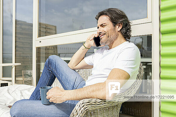Man holding mug talking on smart phone while sitting on sofa in balcony