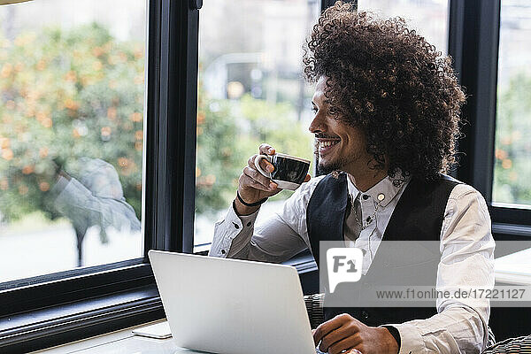 Lächelnder männlicher Unternehmer mit Laptop  der einen Kaffee trinkt und durch das Fenster eines Cafés schaut