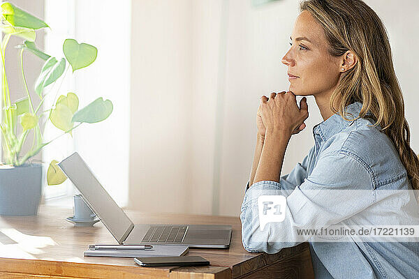 Confident female entrepreneur with hand on chin looking away at home office